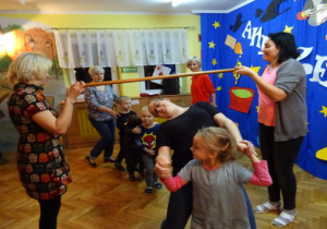 Dzieci i rodzice tańczą przechodząc przez drążek. Dwie nauczycielki trzymają drążek.